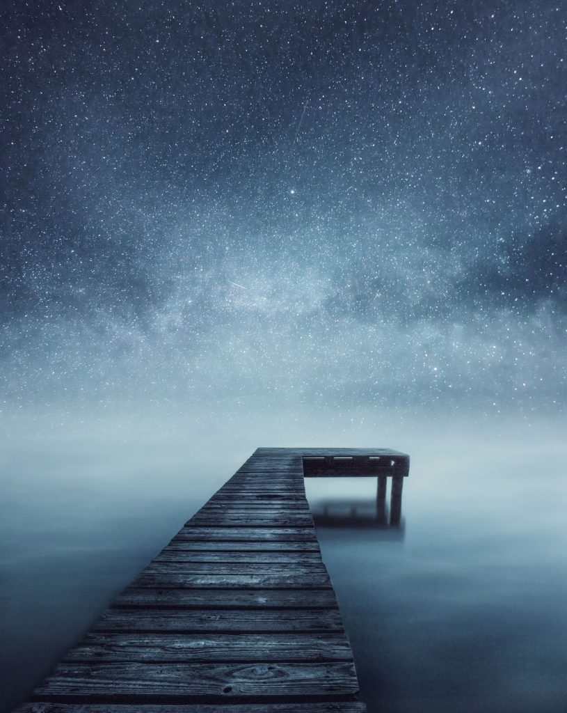 Surreal image of stars at night and a wooden pier over water. 