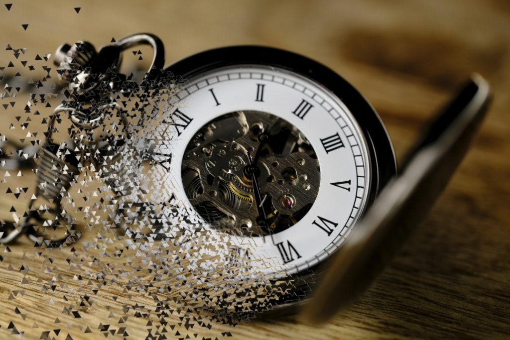 Windup pocket watch landing on a wood grain table and disintegrating, starting in the lower left corner. 