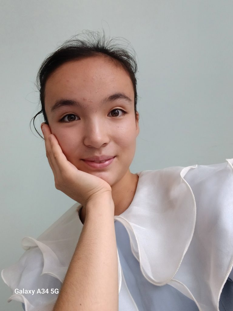 Young Central Asian teen girl with straight dark hair in a ponytail behind her head, her head resting on her hand, and a white ruffly blouse. 