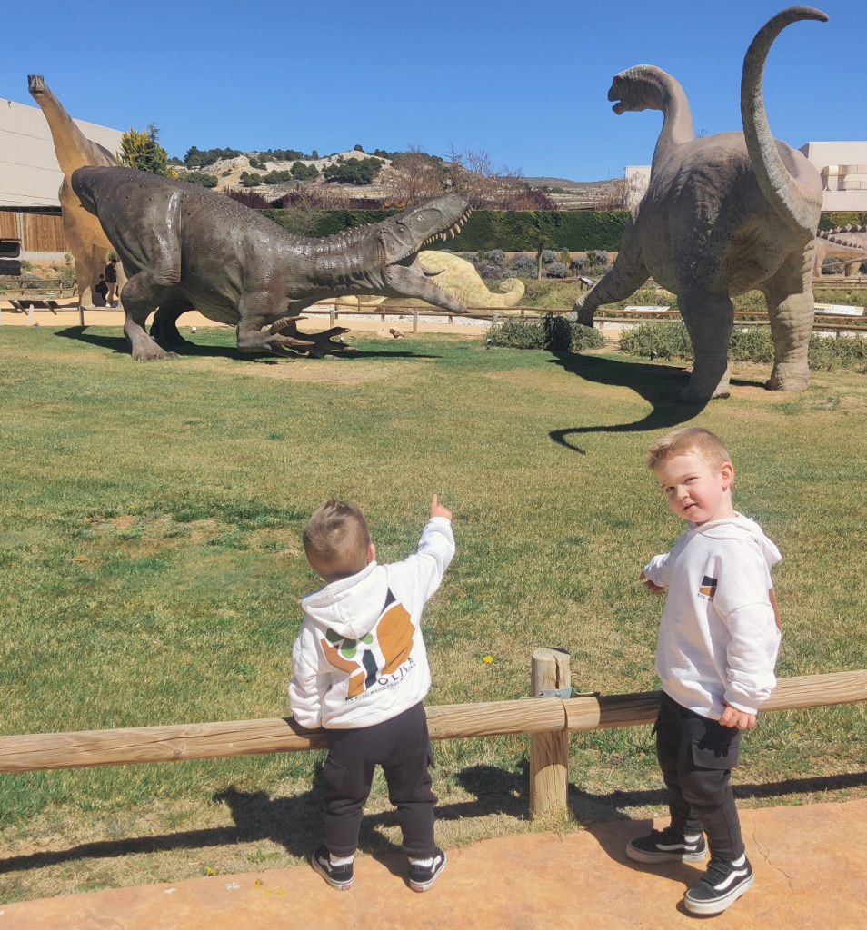 Two preschool age white boys in white sweaters and black jeans and shoes pointing out huge models of dinosaurs out on a field. 