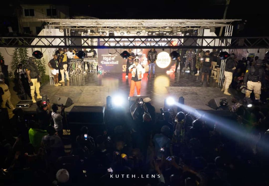 Performer Shatta Wale, in a white jacket, black top, and orange pants, sings on stage in the spotlight to a crowd of fans.