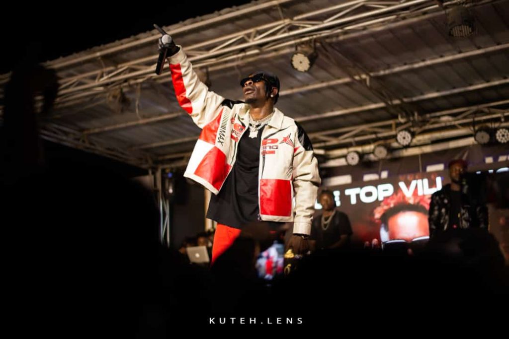 Shatta Wale up close in a red and white jacket and black top holding the microphone.