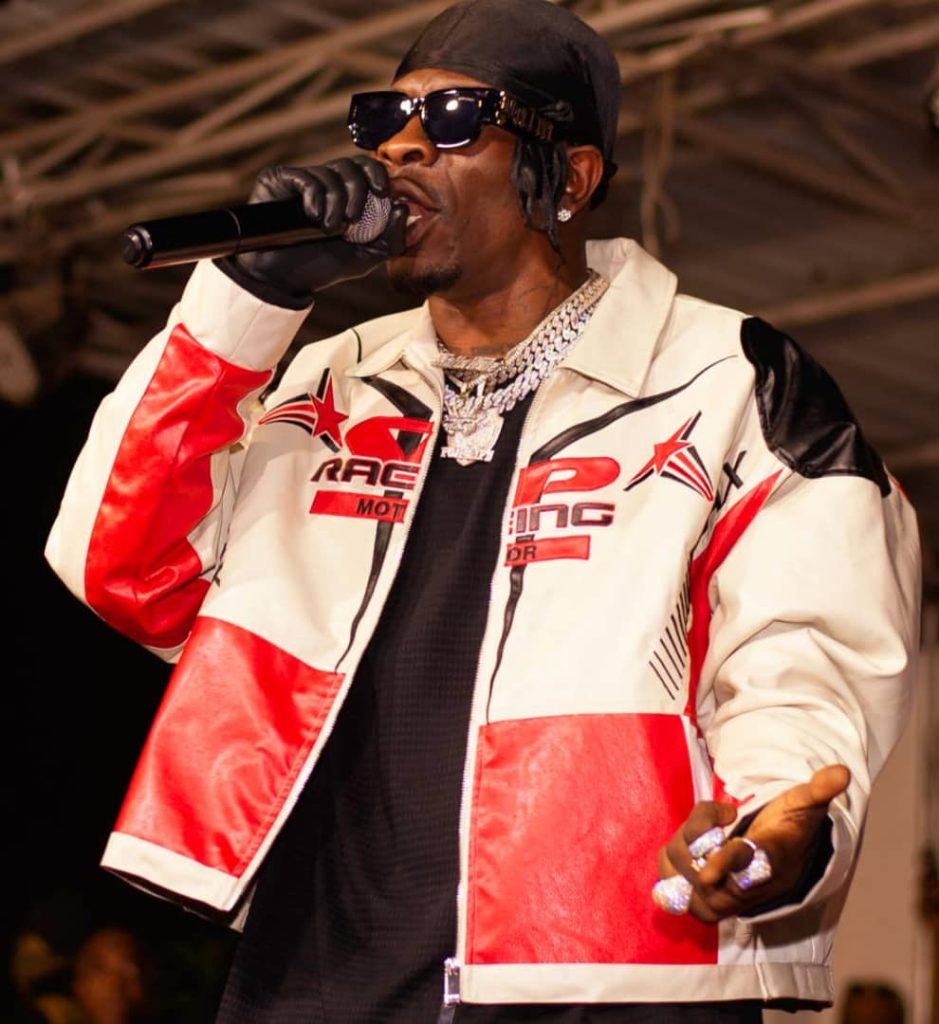 Black man with a black cap, sunglasses, an earring, large gold and silver necklaces, a red and white motorcycle jacket, and a black top sings into a microphone. 