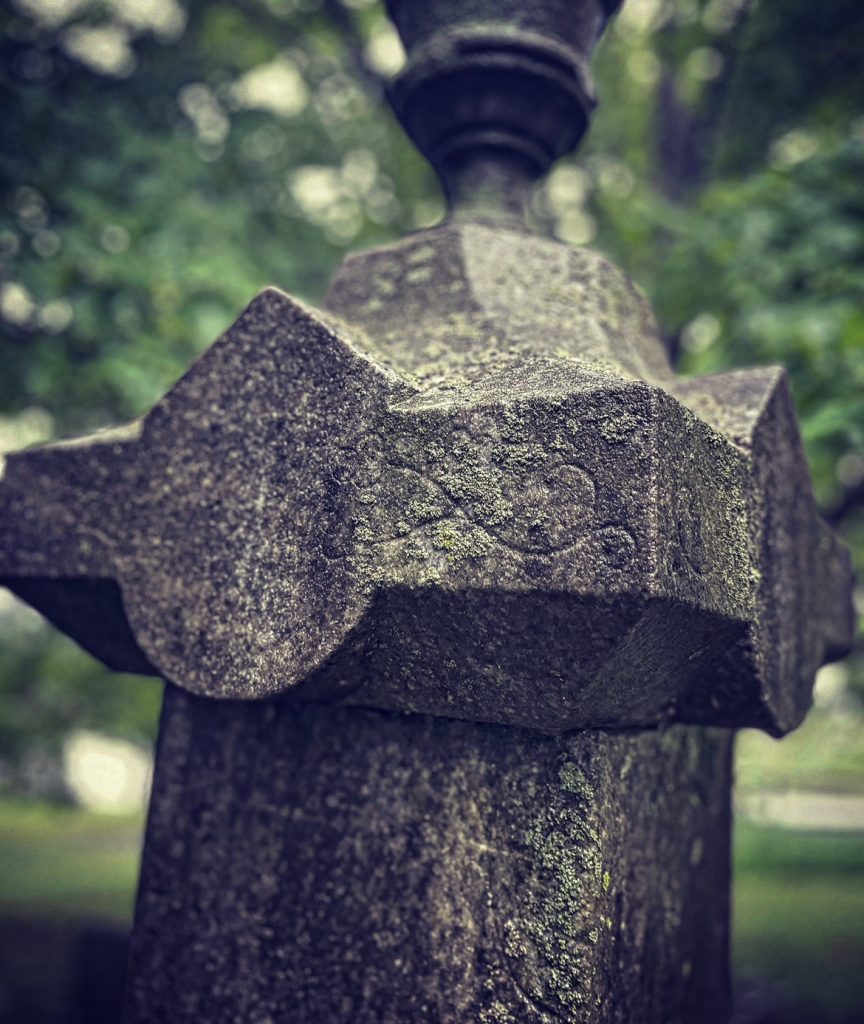 Closeup of a stone post with a filigree design carved into it, with a bunch of trees in the background, out of focus. 