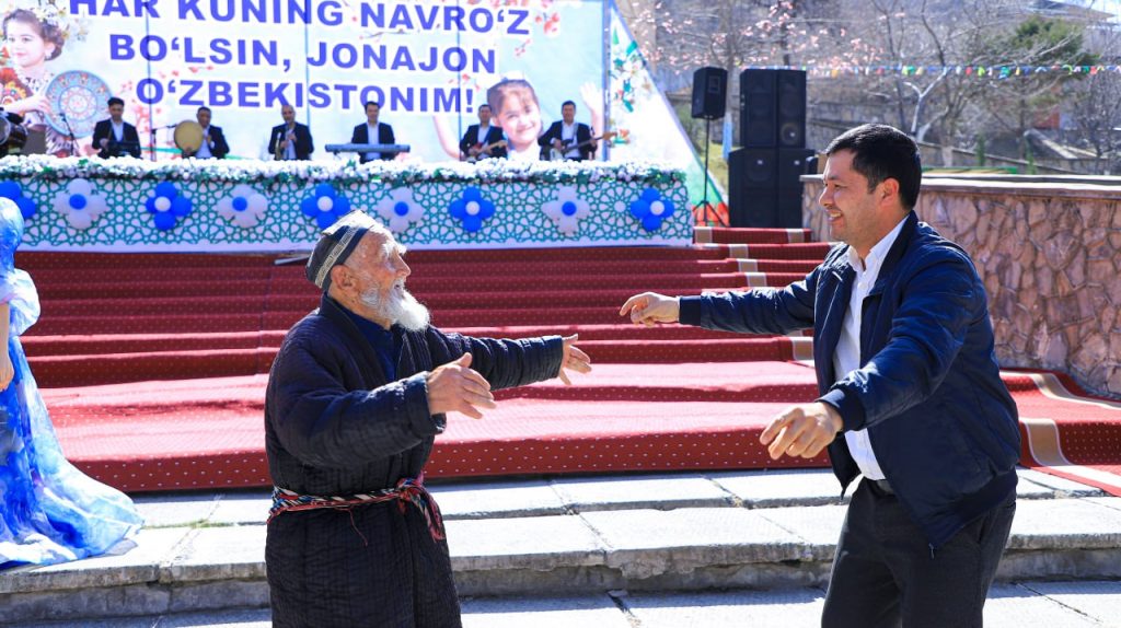 Hamza Khakimzada Niyazi, a young middle aged man with short brown hair and a white collared shirt with a blue jacket, meets and prepares to embrace an old man with a white beard and cape and multicolored string belt and embroidered cap. They're in front of steps with a red carpet and people with microphones at a diplomatic news event. 