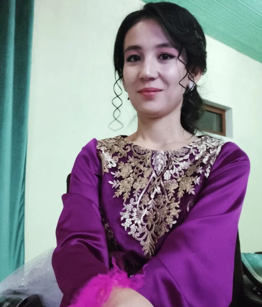 Young Central Asian woman with long black hair up behind her head in a braid and a dark purple silk top with white floral designs at the top. She's outside near a window. 