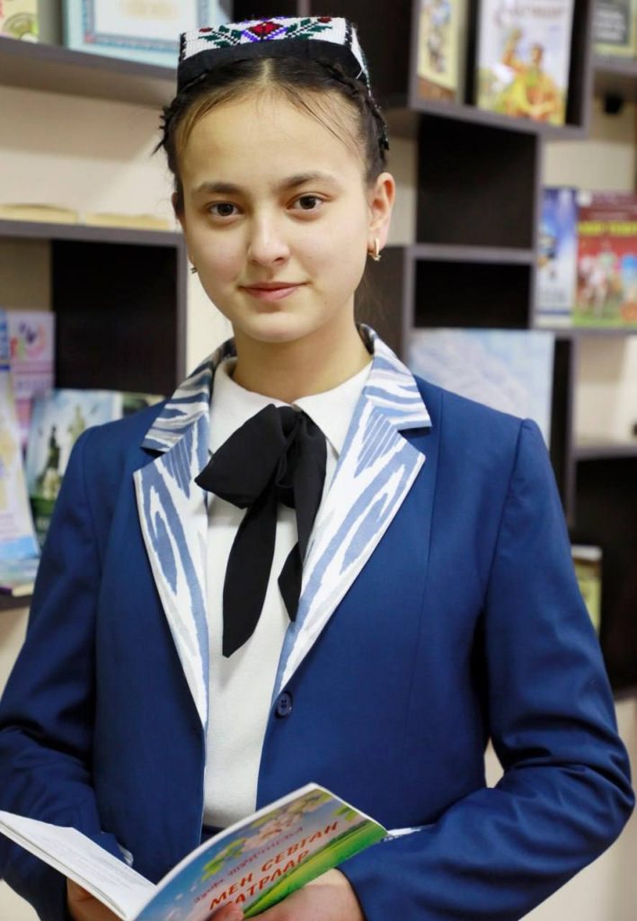 Central Asian teen girl with an embroidered cap on her head with a floral design. She's got brown eyes and straight black hair in a ponytail and earrings and a blue jacket and a white collared shirt with a black bowtie. She's holding and reading a book that looks like a pamphlet or children's book. 