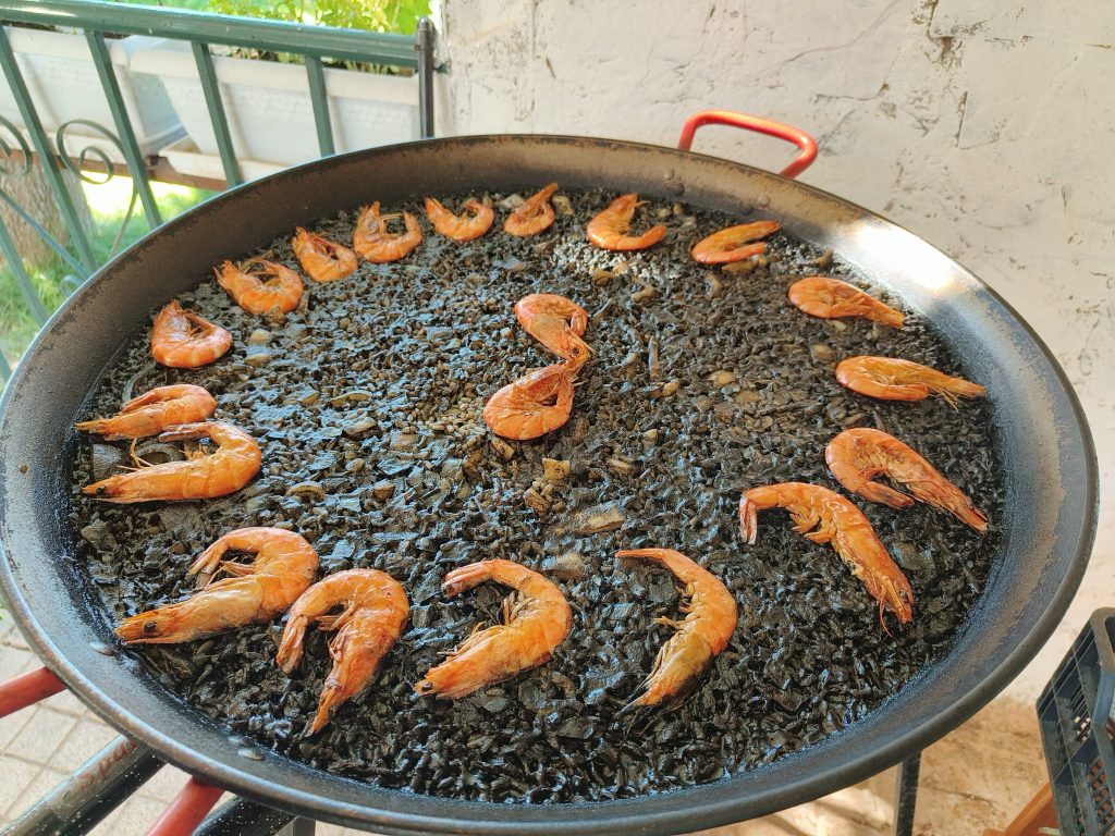 Large pan full of seafood paella, pink shrimp or prawns out on a bowl of black rice. On a balcony on a sunny day of a home. 