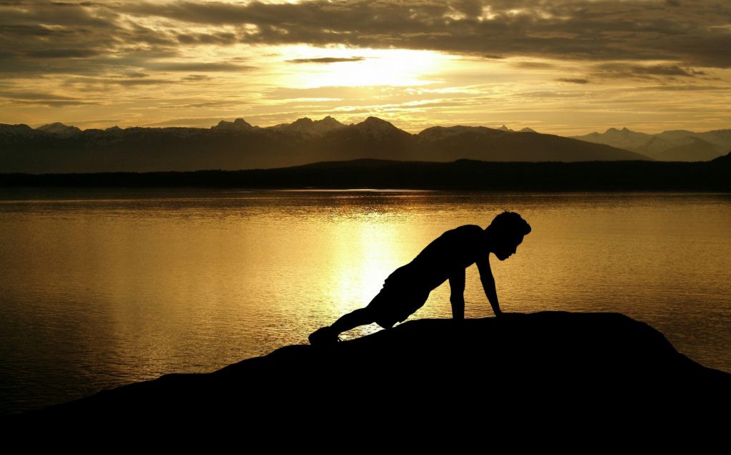 Person doing pushups on a rock by a lake with mountains in the distance and clouds in the sky. Sunrise or sunset. 