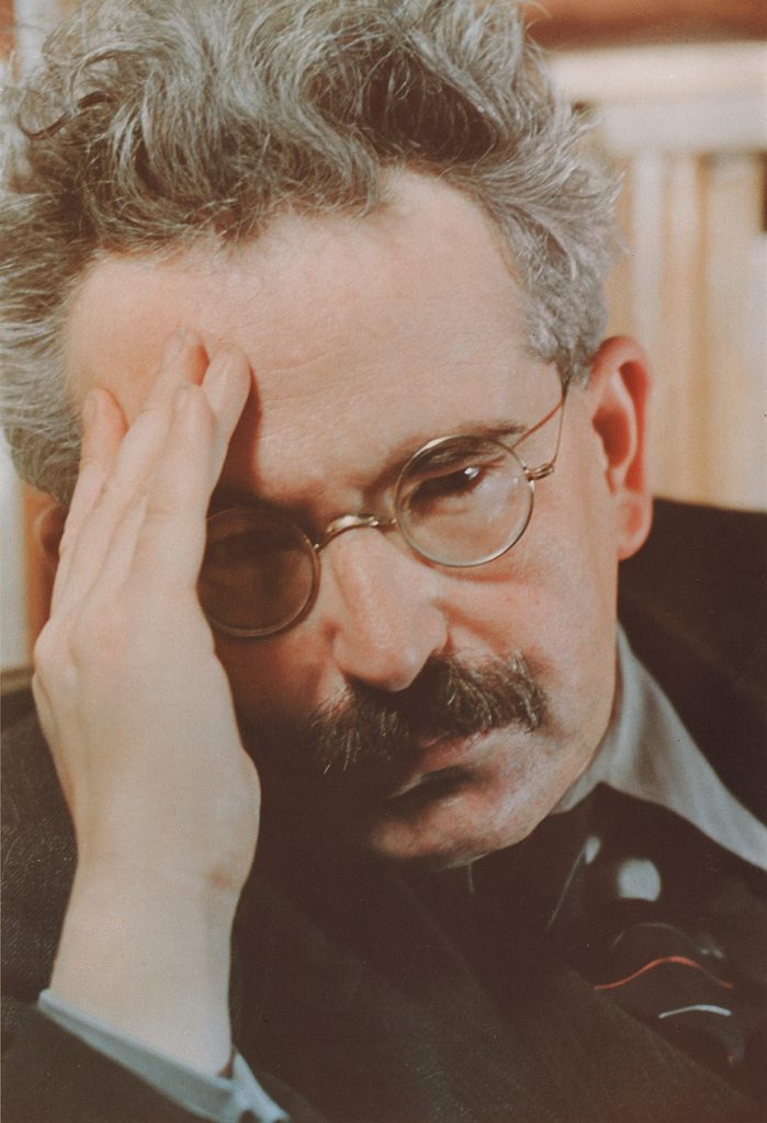 Older white man resting his head on his hand in thought. He's got reading glasses, a suit and tie, and a mustache. 