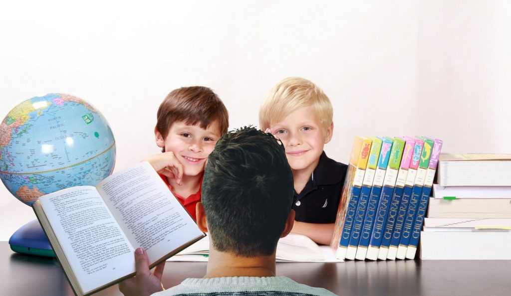 A man of color sits with his back to us and holds a book that he's reading to two white boys, one with blond hair and the other with brown hair. There are encyclopedias and other books and a globe stacked on a table between the man and the boys. 