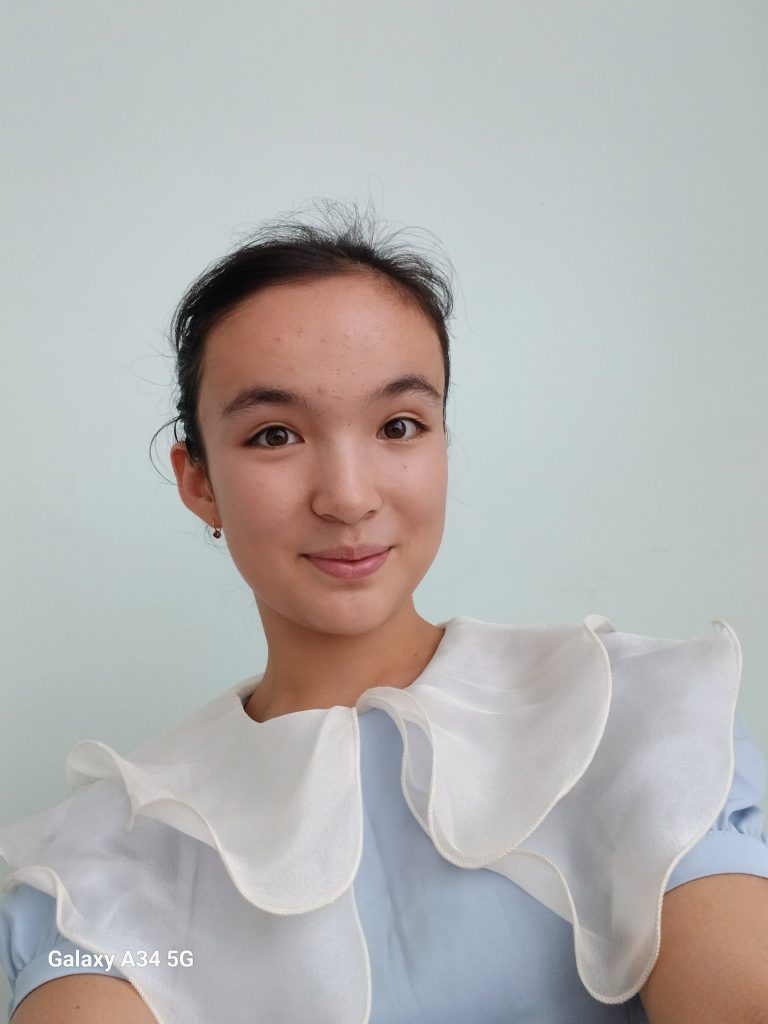 Central Asian teen girl with her hair in a bun behind her head. She's got small earrings and a blue top with white ruffles. 