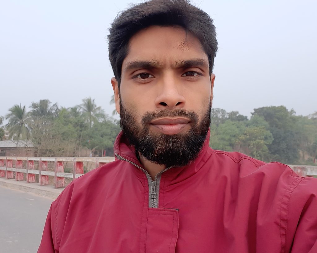 Photo of a young South Asian man with short dark hair and a short trimmed beard and mustache. He's got on a red jacket with a zipper and behind him is a street and barrier and trees. 