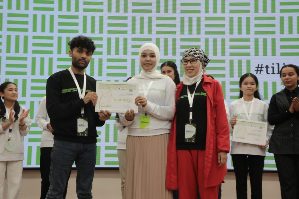 Feruza Axmadjonova stands in the middle, in a white blouse and white headscarf and pink skirt holding a framed certificate together with a darker-skinned guy with a black and green sweater, blue jeans, and dark curly hair. On her right is a woman with a headscarf and a black sweater and a long red coat and red pants. Other students stand nearby, some with their own certificates. Background is white with green lines. 