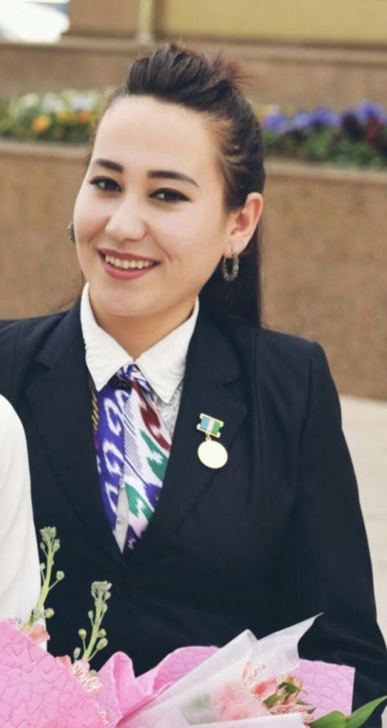Young Central Asian woman with long dark hair, brown eyes, small hoop earrings, standing in front of a planter full of multicolored blooming flowers. She's got on a dark suit and tie and a medal and is holding on to a bouquet of flowers.
