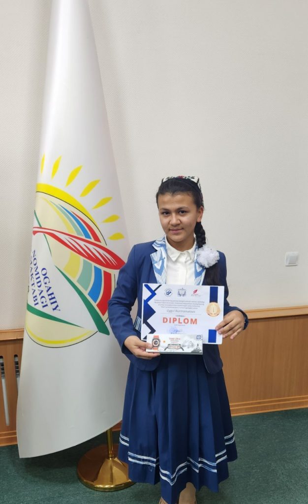 Central Asian teen girl in a long blue jacket and skirt and white collared shirt and an embroidered headscarf holding a diploma and standing in a classroom in front of a banner. 