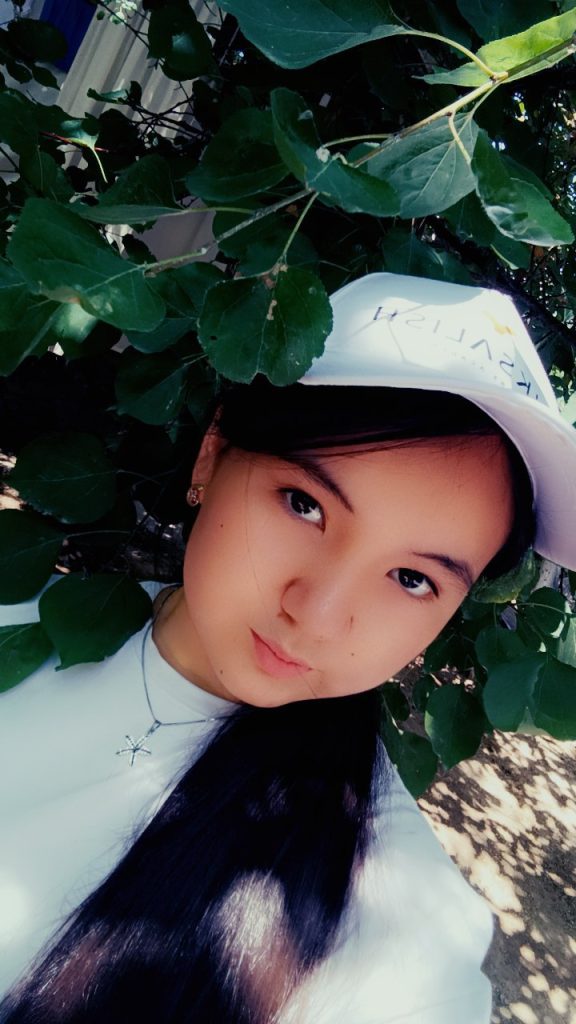 Young Central Asian girl standing in front of a leafy bush at an angle. She's got a white shirt and long dark hair and a white baseball cap. 