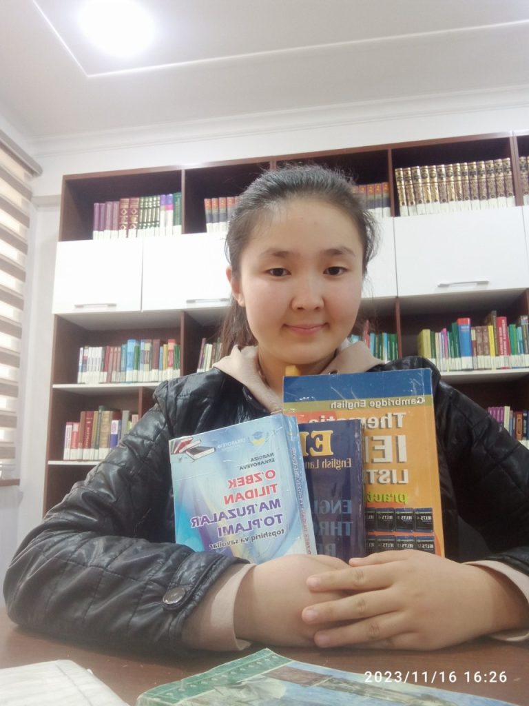 Central Asian teen girl with dark straight hair with a black jacket on and a set of textbooks in her arms. Full bookshelf behind her. 