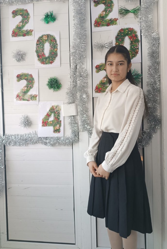 Central Asian teen girl with long dark hair behind her head, a ruffly white blouse, and a long black skirt standing in front of a door decorated by holiday garlands reading 2024. 