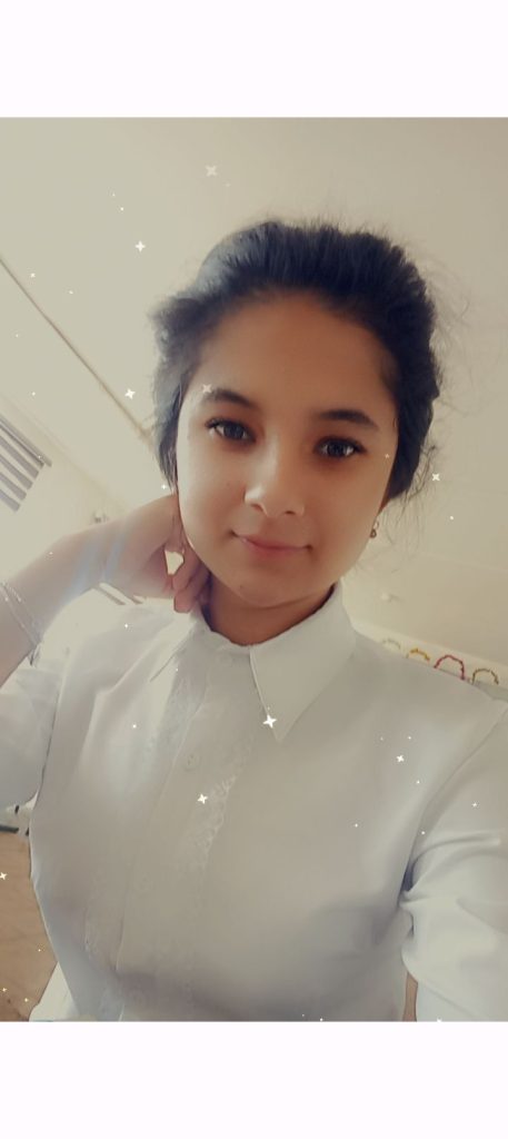 Young Central Asian teen girl with dark straight hair and dark eyes. She's resting her hand on her neck and wearing a white collared blouse. 