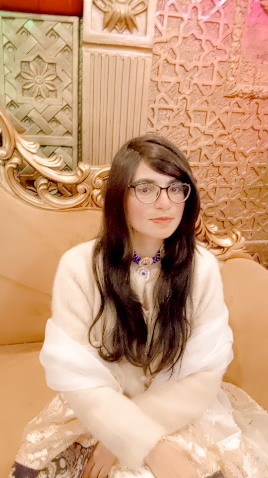 Young light-skinned woman with long dark hair, glasses, a white blouse and blue and gold and white necklace seated on a fancy couch. 
