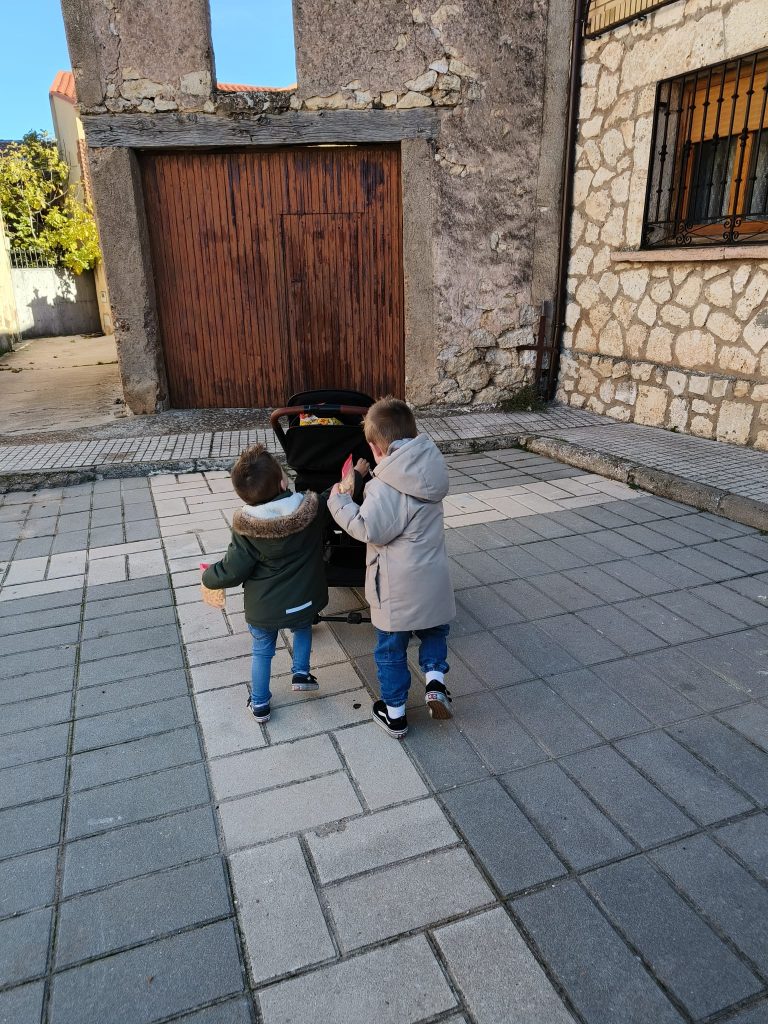 The two boys in thick jackets standing in a concrete path in front of an old stone building with iron bars on a window and a brown door. Sunny day with trees. 