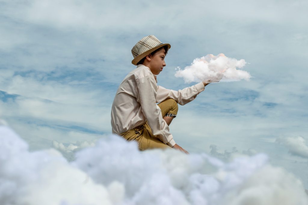 Young boy of indeterminate race in a long sleeve tan shirt and long brown pants and a hat squatting and holding a realistic looking cloud. Other clouds surround him. 