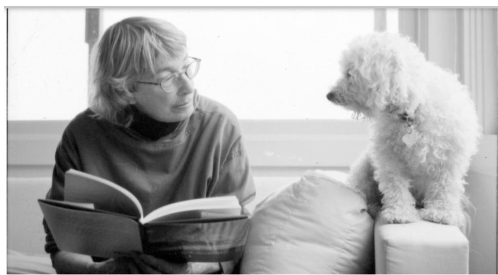 White woman with short light hair and reading glasses and a turtleneck sits on a couch in front of a window holding a book and looking at a fluffy dog 