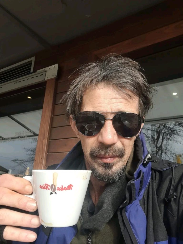 Middle aged white man with short dark hair and dark sunglasses wears a coat and holds a cup of coffee in front of a wood building with windows (a cafe?)