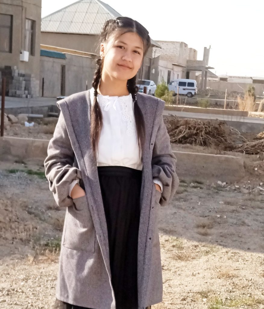 Central Asian teen girl with dark hair in pigtails stands in a construction site i a grey jacket with a white blouse and black skirt. Dirt, dry grass, bricks and vans and buildings are behind her. 