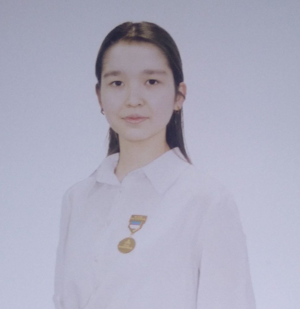 Central Asian teen girl with long dark hair and dark eyes and small earrings wearing a white blouse with a medal on her right breast. 