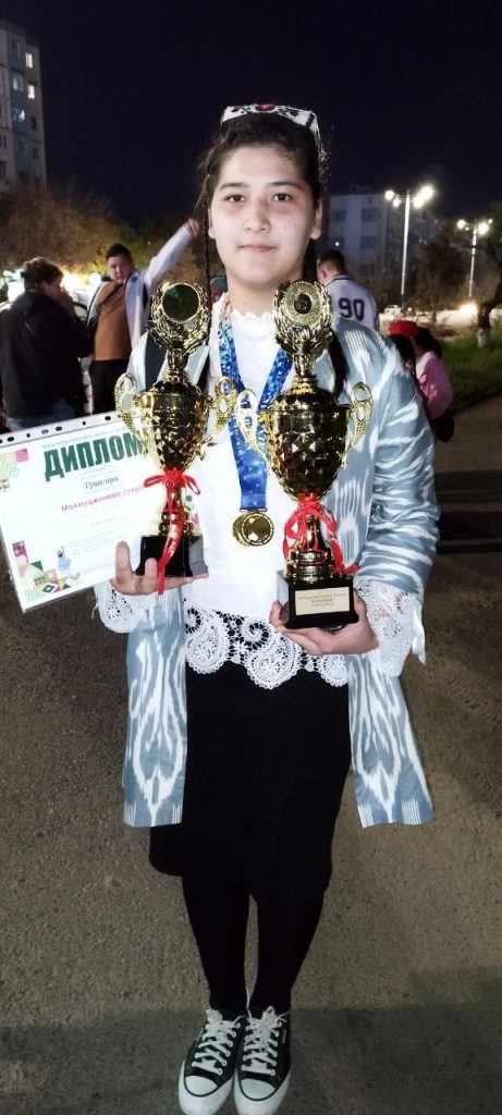 Young Central Asian girl with a woven headdress and long dark hair holding two trophies. Out in the city at night. She's got a silver or blue coat, a white lacy blouse, black pants and tennis shoes.