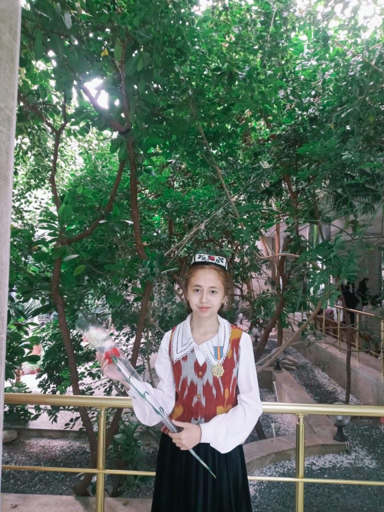 Central Asian teen girl in a brown and orange and gray vest and white blouse and black skirt holds a single red rose and stands in front of a tree. 