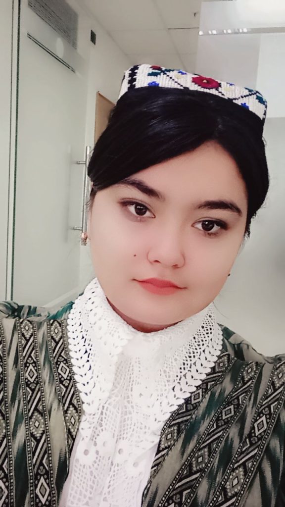 Central Asian teen girl with straight dark hair, brown eyes, small round earrings, a lace collar on a patterned black and gray blouse. 