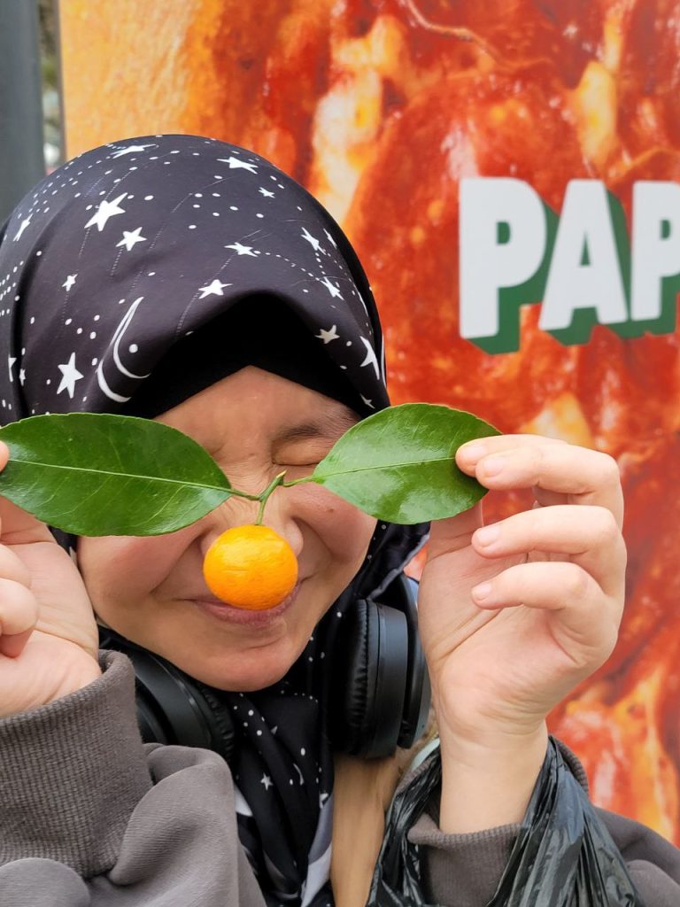 Central Asian young woman with a headscarf with the moon and stars and earphones and a tangerine and two green leaves in front of her face and a gray sweater. 