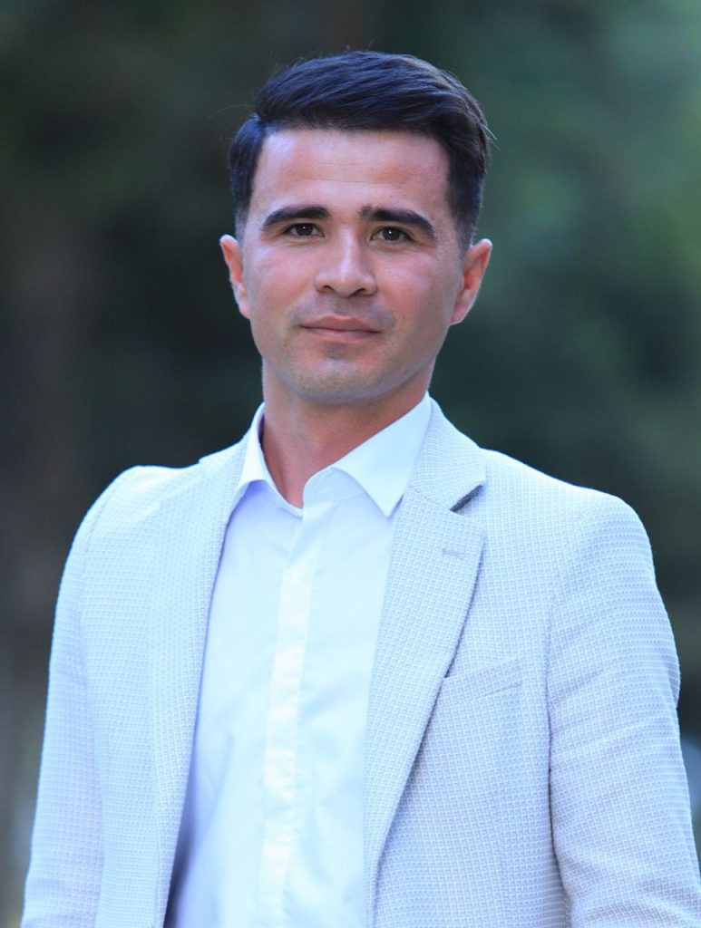 Clean cut young Central Asian man with dark short hair and a white collared shirt and light colored jacket. 