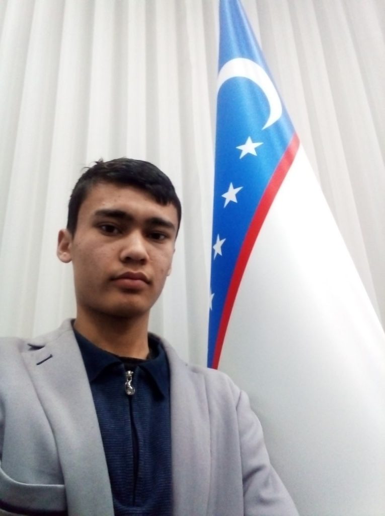 Central Asian teen boy with short brown hair and brown eyes and a gray coat and blue jacket standing on a stage in front of white curtains and the Uzbek flag