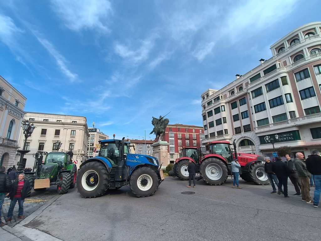 Blue, red, and green tractors parked in the middle of a street with tall buildings and blue sky and clouds and a statue of a winged figure with a sword and old time streetlamps. Burgos, Spain. 