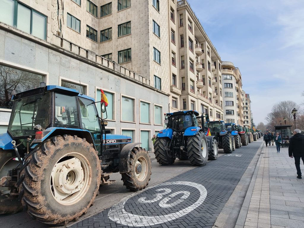 Line of blue tractors with dirty wheels lined upon the sidewalk next to tall buildings in Burgos. People walk down the street. 