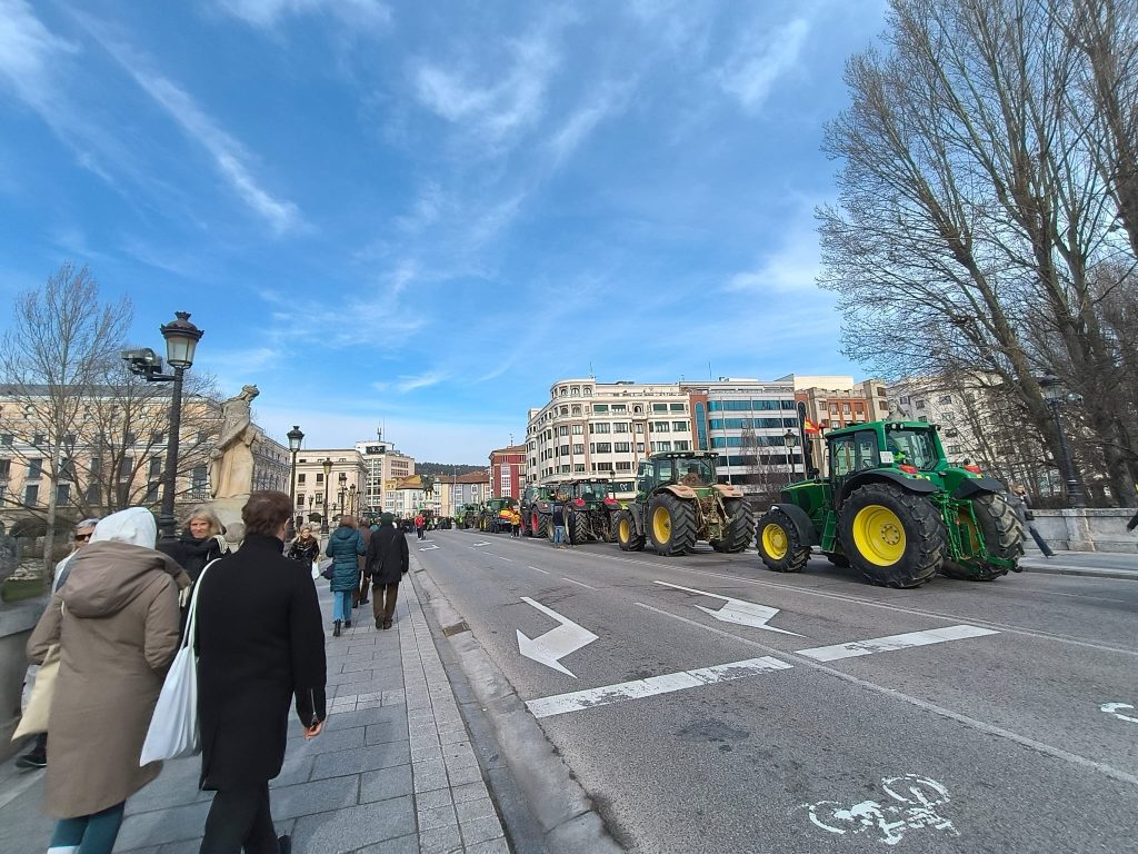 Green tractors parked in a line with barren trees, blue sky, and the city street, lamps, buildings, and a few pedestrians. 