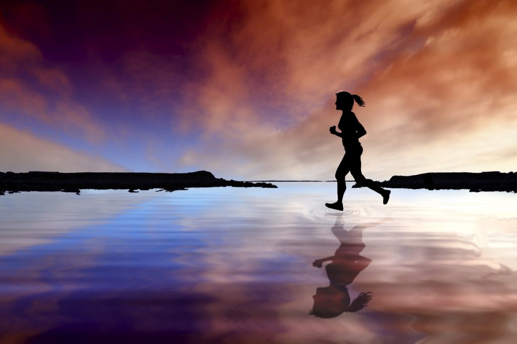 Female shadowed figure of indeterminate race with a ponytail running to the left of a pond between two bits of land among water that reflects the pink and blue and white cloudy sunrise or sunset.