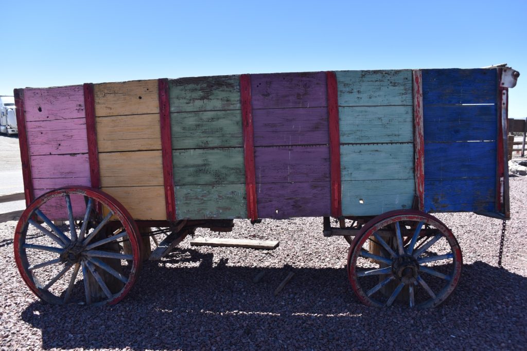Wooden wagon with wooden wheels on gravel. Painted in stripes of blue, purple, green, yellow, and pink. 
