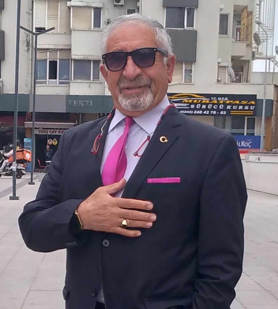 Older light skinned Central Asian man with sunglasses and a suit and a pink tie. He's got a watch and a wedding ring and is standing on a sidewalk in front of an apartment building and storefront. 
