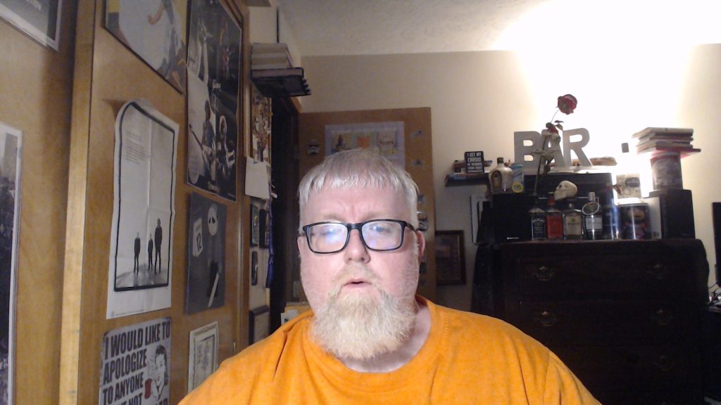 Middle aged white guy with a beard, blonde hair, and reading glasses wearing a big orange tee shirt. He's in a room in front of a wall full of posters and a nightstand with cologne bottles and a rose. 