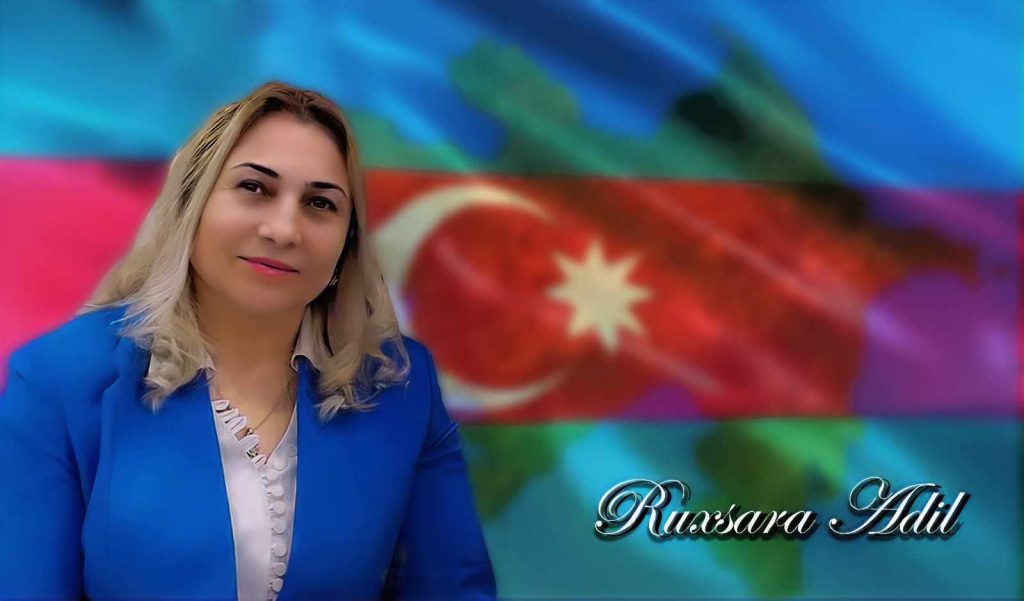 Light skinned woman standing in front of the Turkish crescent and star flag in a red stripe across a blue and green banner. She's got curly blonde hair and a blue jacket over a ruffly white blouse. 