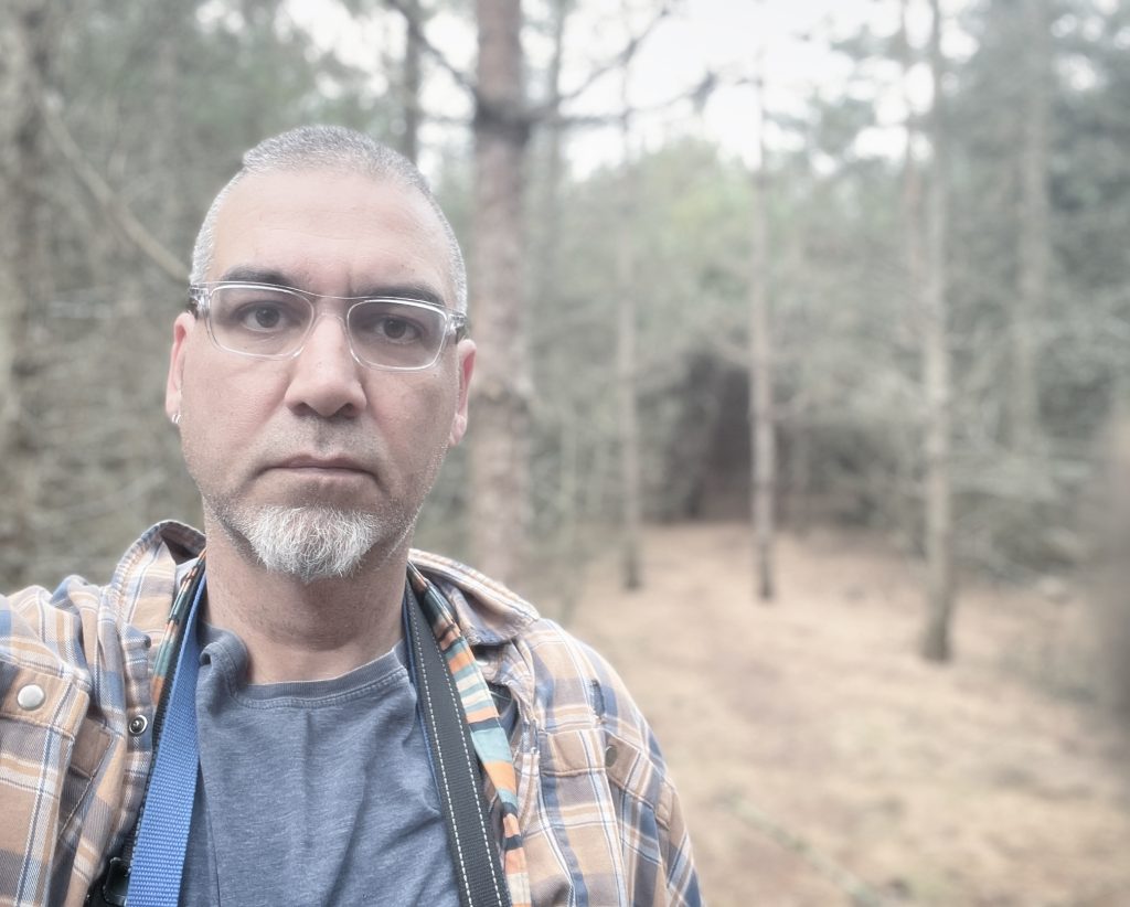 Middle aged white guy with a trimmed beard and reading glasses with a plaid coat over a gray tee shirt stands in a clearing in a forest. 