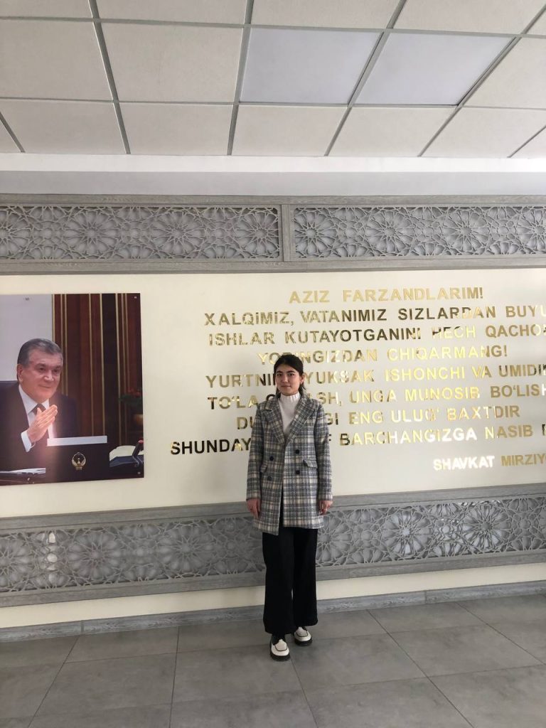 Central Asian teen girl in a woolen gray and white jacket and white sweater and black pants standing in front of a wall with a quote in gold lettering and filigree designs and the photo of an important white man in a suit holding up his hand. 