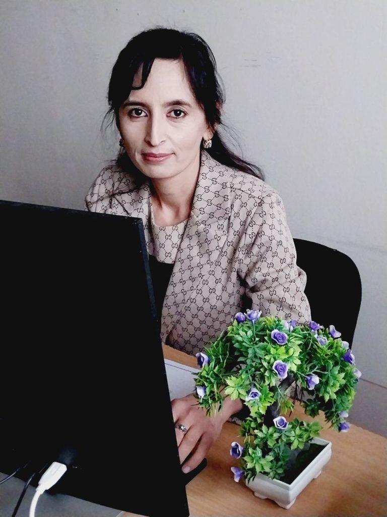 Central Asian woman seated at a computer desk with a light pink patterned jacket, black blouse, long dark hair and earrings, and a flowering plant at her side. 