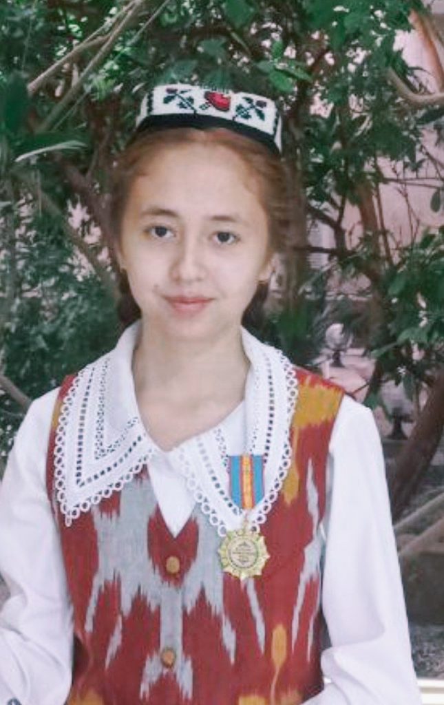 Young teen Central Asian girl with an embroidered headdress, brown hair in a braid, brown eyes, and a lacy white top with a red and orange vest. She's standing in front of a tree. 