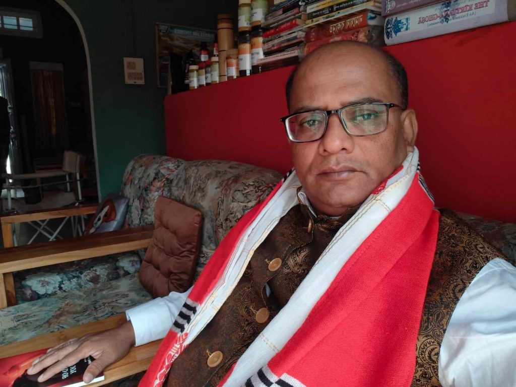 Middle aged South Asian man with reading glasses, a red shawl and brown patterned and buttoned vest over a white shirt. He's sitting on a couch with some books on a shelf behind him. 
